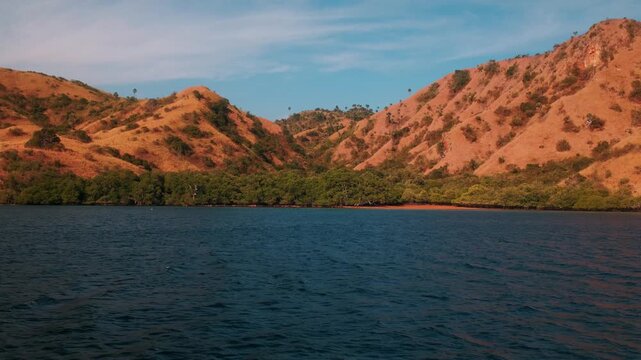 View of Pulau Kalong Rinca with its distinctive dry, golden-brown hills rising above the deep blue waters of the bay under a clear sky in Komodo, East Nusa Tenggara, Indonesia.