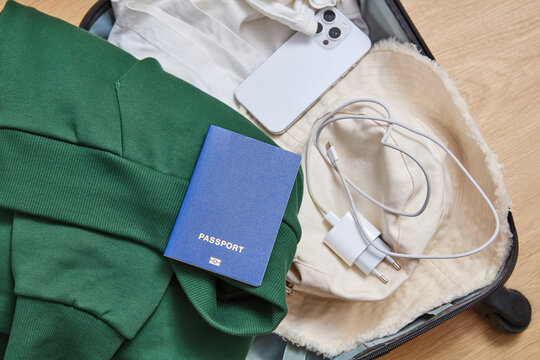 Open suitcase packed with clothes, smartphone, charger, hat and blue passport for travel