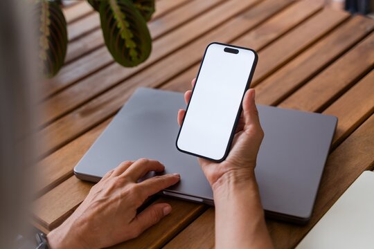 Hand holding smartphone with blank screen over laptop on wooden table