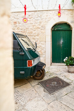 Dark green Piaggio Ape 50 parked in front of a green wooden door in a whitewashed old town alley, classic Italian three-wheeler, Ostuni, Puglia, southern Italy 27.11.2025