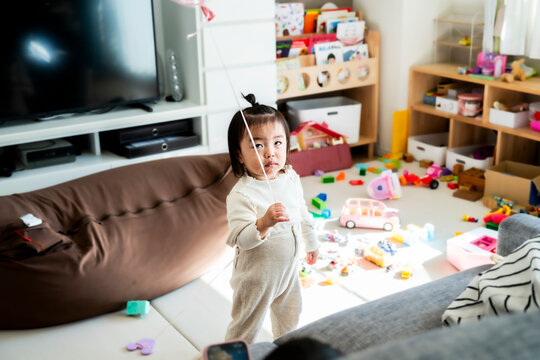 Cute Asian Toddler Girl Standing in Messy Playroom Holding Balloon String at Home