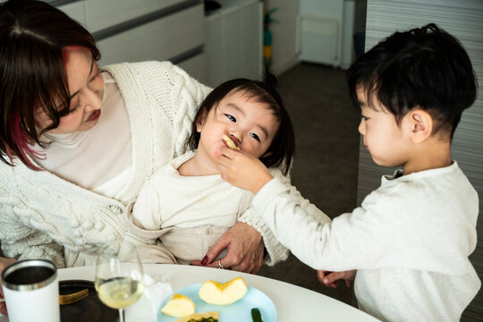 Kind Asian Boy Feeding Food to Toddler Sister Held by Mother at Table