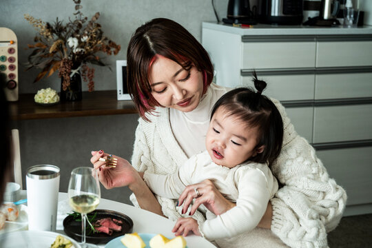 Asian Mother Holding Toddler Girl Sitting at Dining Table During Home Meal