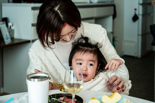 Asian Mother Holding Toddler Girl Sitting at Dining Table During Home Meal
