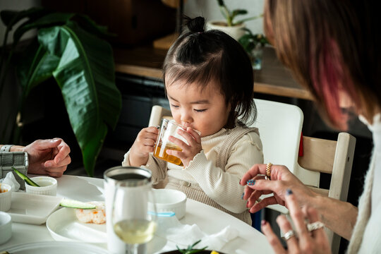 Cute Asian Toddler Girl Drinking Tea from Glass Cup at Dining Table with Family