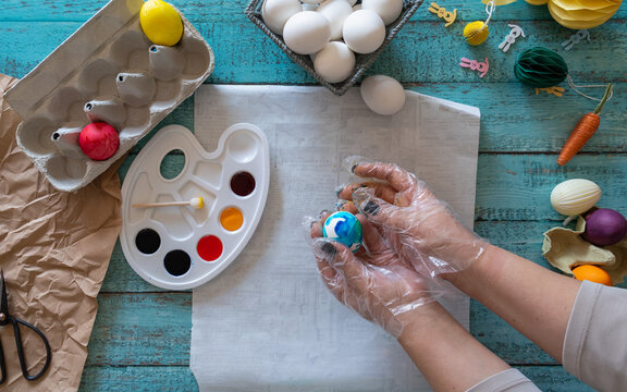Creative Easter craft scene showing gloved hands painting decorative eggs with a watercolor palette surrounded by additional eggs and colorful craft supplies on a rustic tabletop