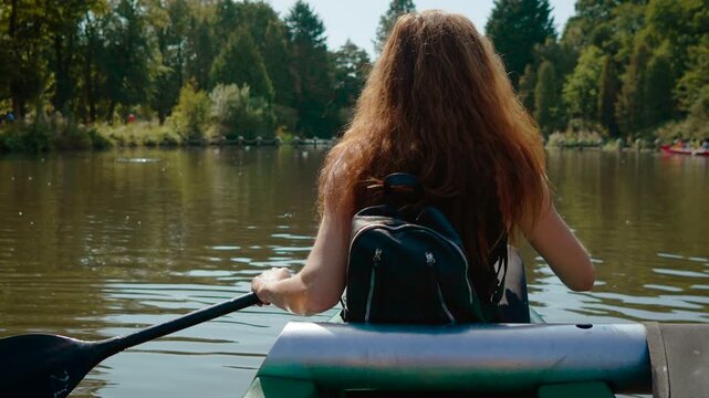 Young woman paddling canoe across calm lake during sunny summer day. Outdoor recreation in green park with backpack and paddle. Active lifestyle, kayaking adventure and nature travel