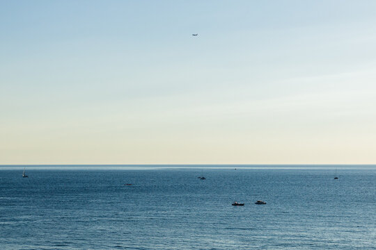 Aerial view of a calm ocean with several boats floating on the water, a distant airplane flying in the clear sky above the horizon during daylight