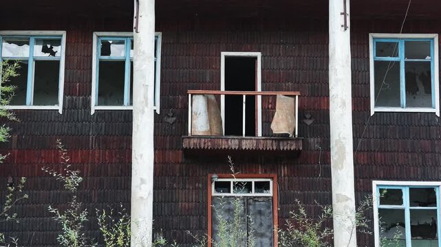 Static view of a weathered wooden building featuring broken windows, a rusted balcony, and overgrown vegetation, maintaining a consistent unmoving perspective of the deteriorating structure.