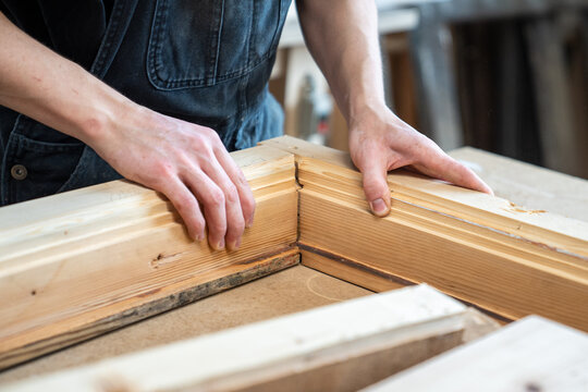 Carpenter assembling wooden parts of vintage window frame at workbench in restoration workshop, craftsman putting together details of wooden frame at workplace, checking fitting and adjusting angles.