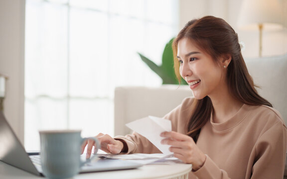 Smiling Asian woman checks receipts and household expenses while using a laptop at home, representing budgeting, financial planning, bill payment, money management and personal finance.