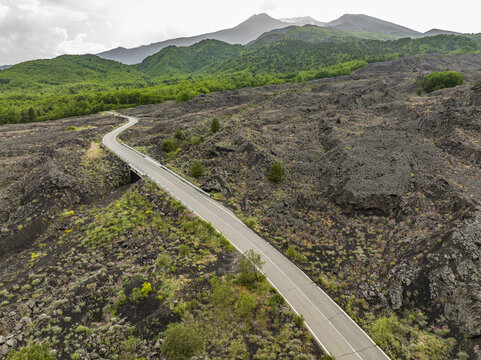 Aerial view of a winding road crossing through the volcanic landscape of Mount Etna with black lava rocks, sparse green vegetation, and distant mountains in Catania, Sicily, Italy.