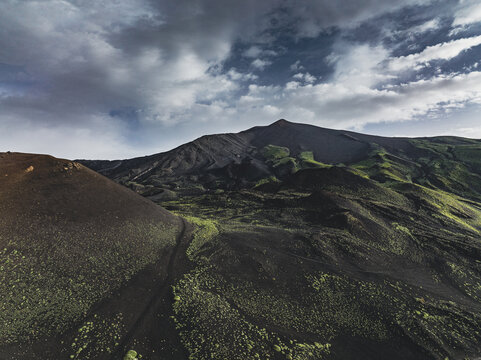 Aerial view of the volcanic landscape of Mount Etna with dark lava fields and patches of green vegetation under a dramatic cloudy sky in Catania, Sicily, Italy.