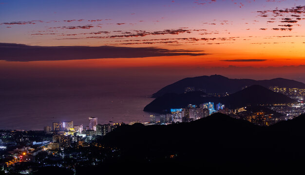 Hilly coastline silhouetted at sunset with lit seaside town Sanya, Hainan Island, China