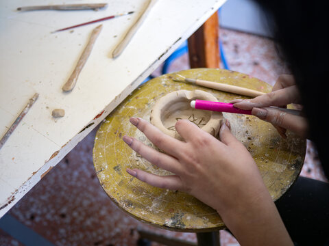 Detail of a person adding decorative elements to a handmade ceramic piece in a cozy art studio. The potter uses a specialized sculpting tool to work on the small clay object resting on a wheel.