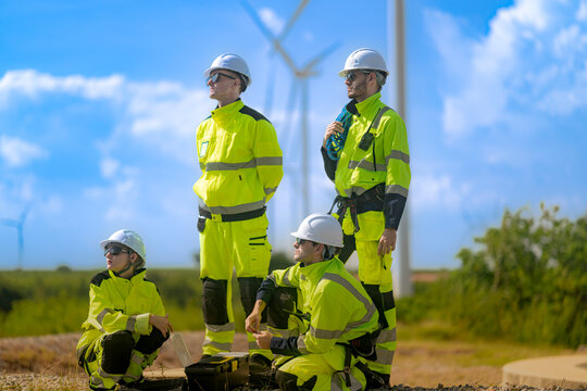Crouching technician opens a black flight case on gravel while two colleagues stand over him with their eyes on the sky and a woman kneels at the left of frame, a breezy day of fieldwork