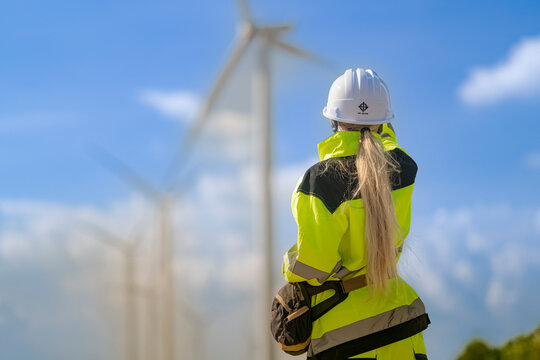Blonde ponytail and yellow high visibility jacket seen from behind as a wind technician looks out at a slowly turning rotor above the flat green landscape, green power generation work
