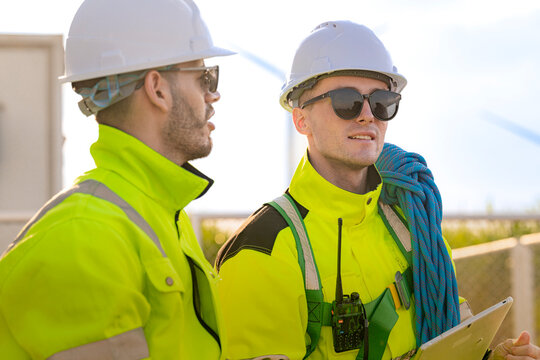 A close two-shot of site colleagues in yellow hi-vis and white hard hats, the younger man smiling faintly as both keep their attention on the far end of the yard