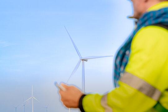 A technician's hand grips a slim stylus at the lower left while a single wind turbine rises through thin cloud, framed by the reflective bands of his yellow jacket