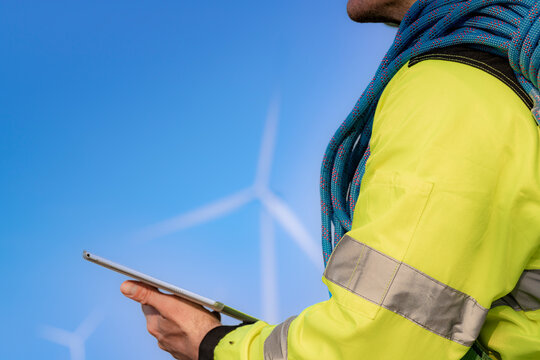 Seen from behind, a technician in a yellow jacket and blue rope coil holds a tablet out at waist height while wind turbines fade into blue haze on the plain in front
