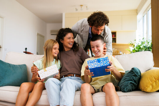 Happy family celebrating an occasion, parents giving present boxes to their smiling children sitting together on a comfortable sofa in a bright living room at home