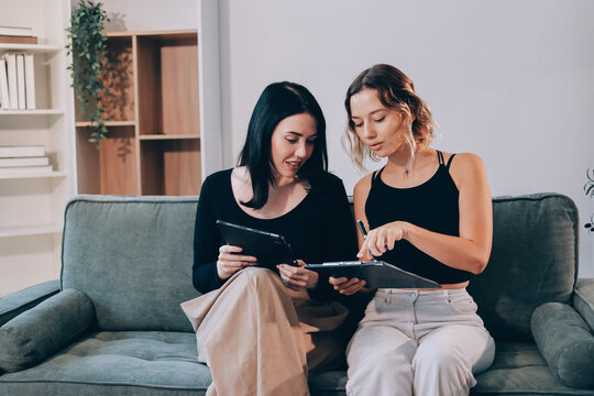 Two young female students using a laptop while sitting on a sofa at home. Student life concept with studying, teamwork, and online learning.