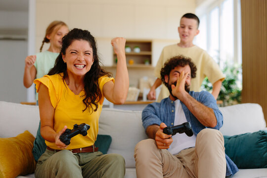 Happy family enjoying a competitive game at home, with the mother cheering her victory and the father reacting to his defeat while children watch and react in the background