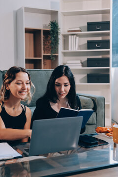 Two young female students using a laptop while sitting on a sofa at home. Student life concept with studying, teamwork, and online learning.