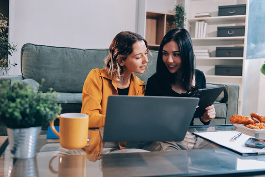 Two young female students using a laptop while sitting on a sofa at home. Student life concept with studying, teamwork, and online learning.