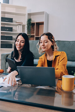 Two young female students using a laptop while sitting on a sofa at home. Student life concept with studying, teamwork, and online learning.