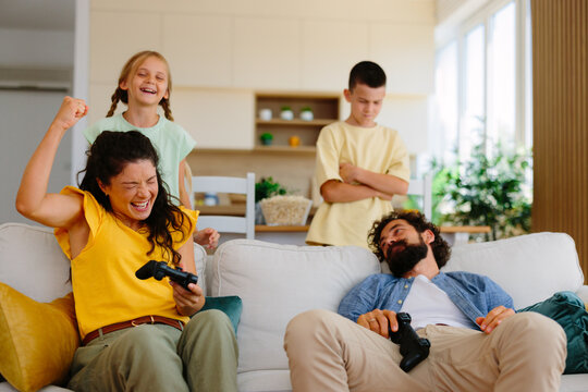 Family members expressing different emotions while playing video games together, with a winning mother and daughter celebrating as the father and son experience defeat in their living room