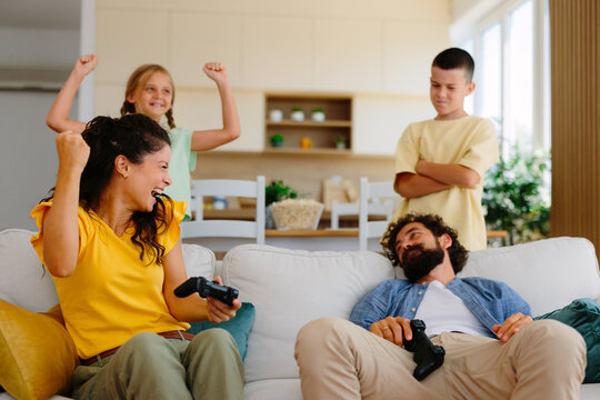 Cheerful mother and daughter celebrating winning a video game match, while father rests defeated on the sofa and son looks disappointed from behind them in the living room