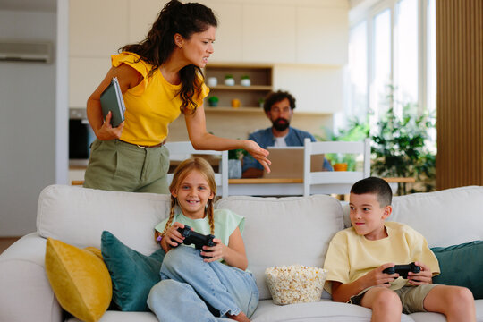 Frustrated mother scolding kids playing video games on the couch with popcorn while father works on his laptop in the background, highlighting screen-time conflict at home
