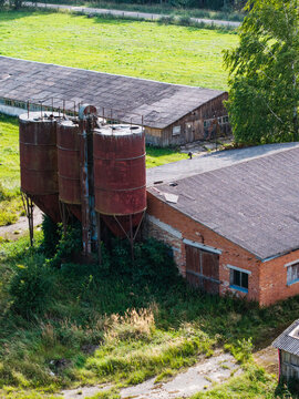 Aerial view of an abandoned Soviet-era agricultural complex with rusted silos and dilapidated brick buildings surrounded by overgrown fields in Kulciems, Talsi Municipality, Latvia.