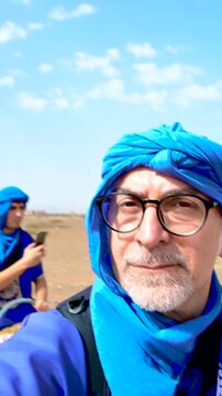 Marrakech, morocco. August 9, 2026. Mature man with glasses and a blue tuareg turban taking a selfie while riding a camel. A caravan of tourists is visible behind him