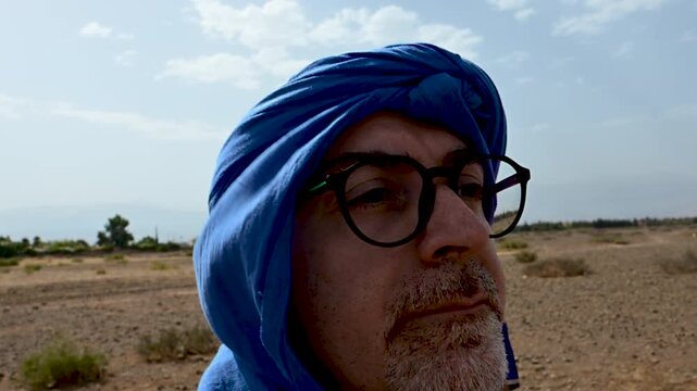 Marrakech, morocco. August 9, 2026. Caucasian man with a beard and glasses wearing a traditional blue turban while taking a selfie on a camel ride in the agafay desert, morocco