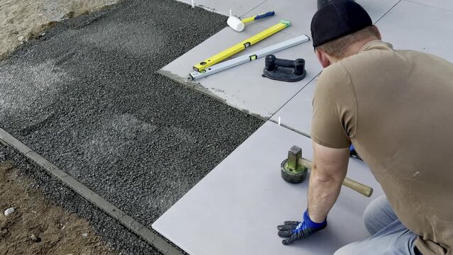 Male worker in gray shirt and black cap lays large tiles on gravel surface, using a rubber mallet and level tool for precise alignment in outdoor setting