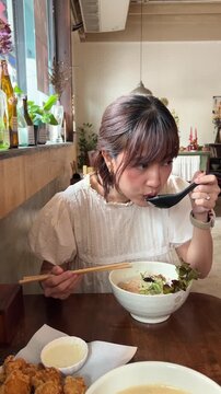 Happy delighted young asian woman eating delicious hot tasty ramen noodle soup with chopstick and spoon inside cozy dining restaurant lunch meal