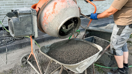 Construction worker using concrete mixer to pour gravel into wheelbarrow on building site, with...