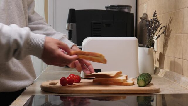 Person's hands placing golden brown toasted bread slices into a white toaster on a kitchen counter with fresh tomatoes, cucumber, and plain bread for a morning meal