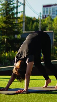 Woman practicing outdoor yoga in downward dog pose on green grass mat surrounded by trees and sunlight during peaceful mindful fitness stretching routine