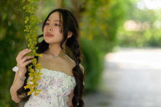 Elegant woman with long dark hair and floral dress standing outdoors near yellow flowering plant