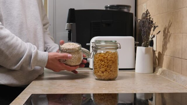 Person's hands holding an open glass jar filled with grains, standing next to another sealed jar of pasta on a kitchen countertop with a coffee machine and toaster