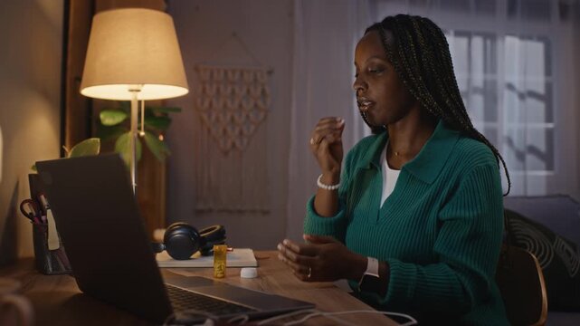 Young adult African American woman sitting at desk at night, taking medication from pill bottle with water while working on laptop. Perfect for mental health and digital burnout campaigns
