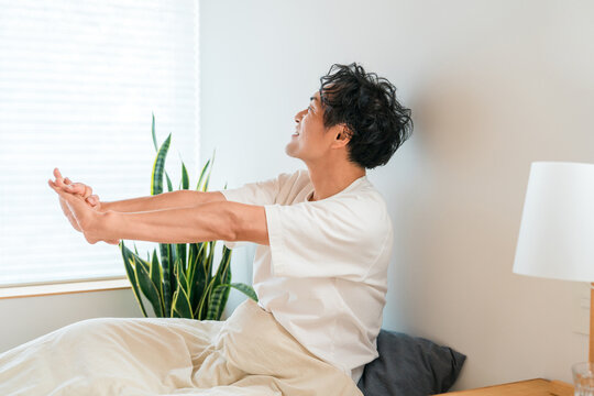 朝起きてベッドの上で気持ちよく伸び,ストレッチをするアジア人中年男性