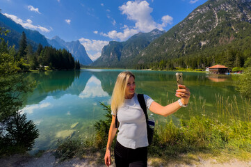 Naklejka premium Young woman with backpack on the lake in alpine mountains, blue sky with clouds, reflection in clear water, at sunset. Summer travel in Dolomites, Italy. Girl on the lake. Hiking 