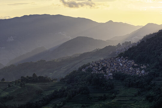Aerial view of a village in the terraced hills of Yuanyang County, illuminated by golden sunset light with lush green slopes in Honghe Hani and Yi Autonomous Prefecture, Yunnan, China.