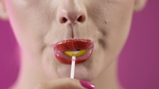 Close up shot of young woman with red glossy lips enjoying a sweet cherry lollipop on vibrant purple background.