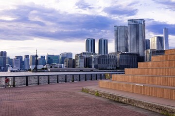 海沿いの公園にあるウッドデッキベンチと対岸に広がる高層ビル群の都市風景（横構図） © Riz