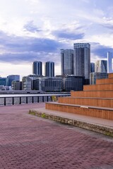 海沿いの公園にあるウッドデッキベンチと対岸に広がる高層ビル群の都市風景（縦構図） © Riz